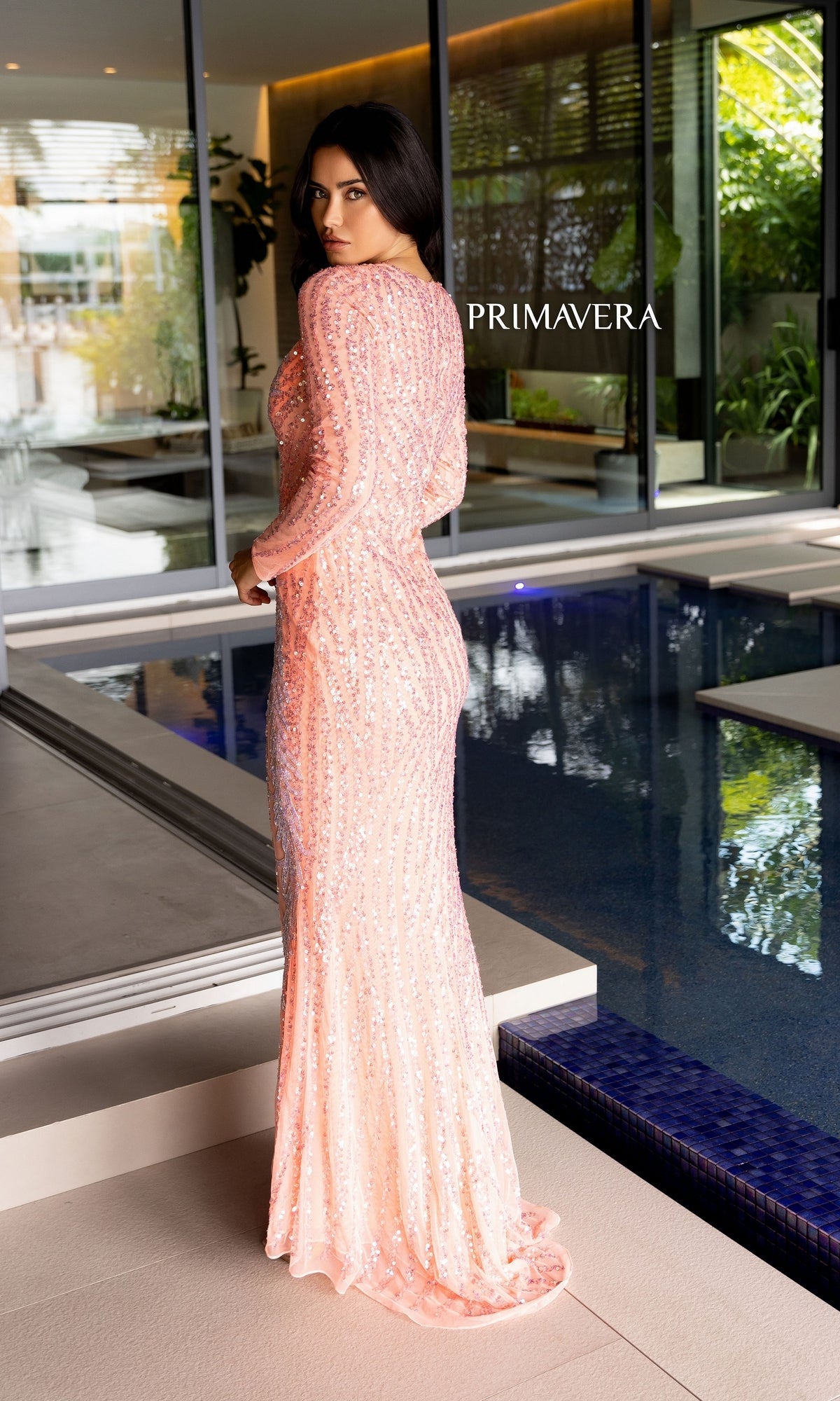 A woman wearing the Long Prom Dress 12117 by Primavera, a fitted light pink beaded formal gown, stands by an indoor pool looking over her shoulder. The brand name